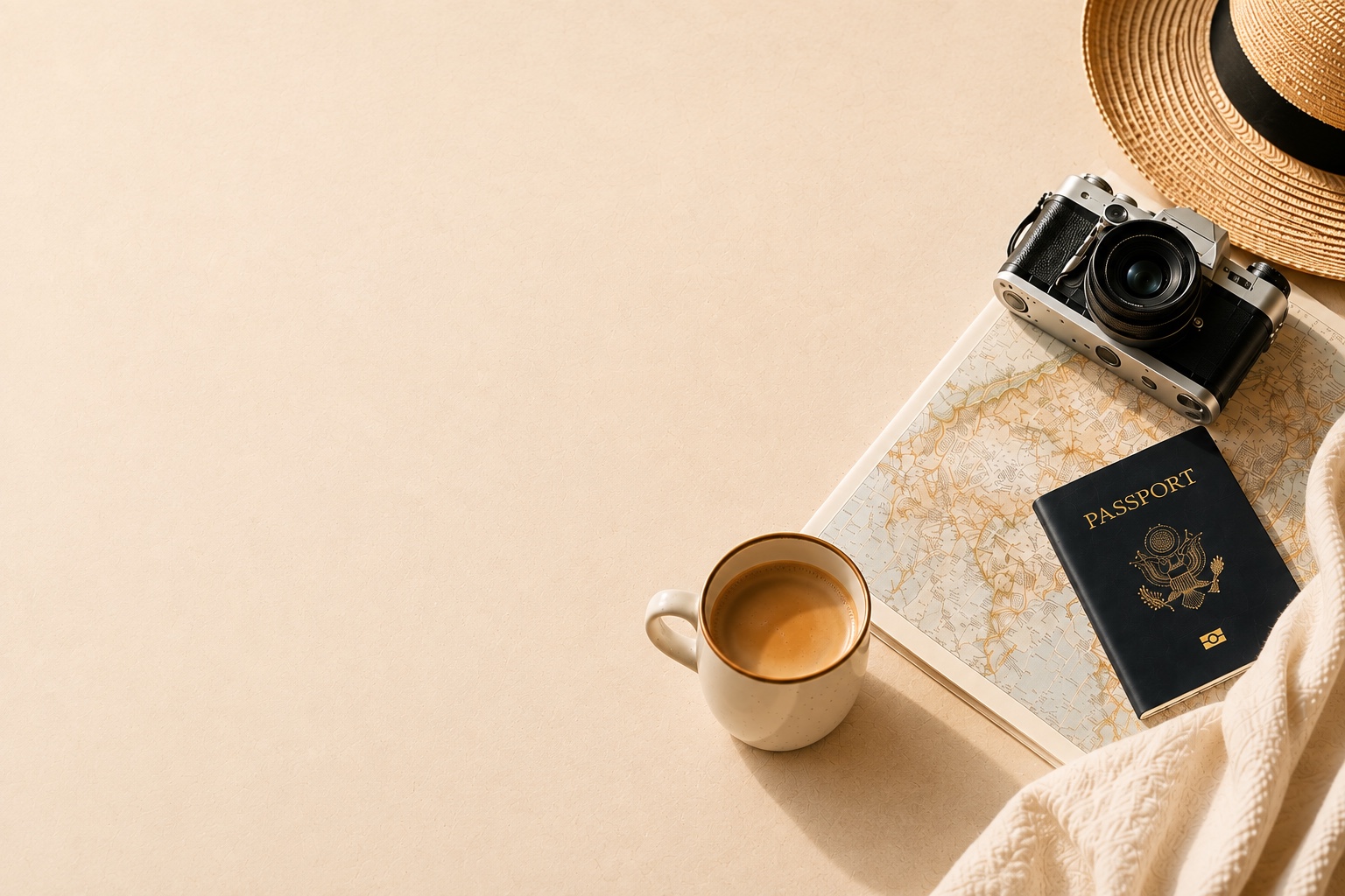 A flat-lay travel still life: a vintage camera, a straw sun hat, a passport, a folded map, and a cup of coffee arranged on a sunlit surface